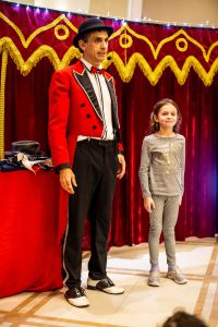 Em um palco, um homem vestido de mágico está ao lado de uma menina