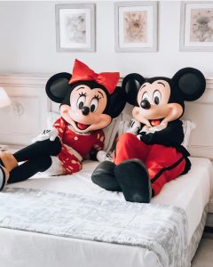 Duas pessoas, fantasiadas de Minney e Mickey, sentadas lado a lado em uma cama do hotel