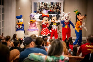 equipe de recreação composta por 6 membros, todos fantasiados de personagens da Disney. Em ordem: pato Donalds, Maragrida, Miney, Mickey, Coelho branco e Pateta. Eles se localizam em um palco e, logo à frente, há inúmeras crianças e pais assistindo ao evento.